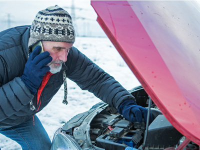 Older man wearing dark gray jacket, hat and gloves on the phone and looking under the hood or a red vehicle